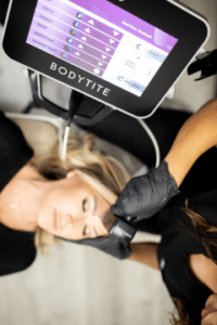 A person receiving a facial treatment using BodyTite equipment. A healthcare professional wearing black gloves is applying the device to the person's forehead. The BodyTite machine's screen displays treatment settings. The person receiving the treatment has their eyes closed and appears relaxed.