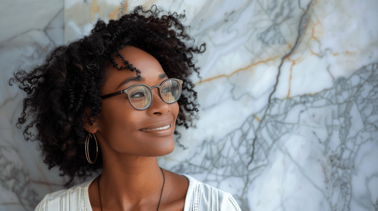 A woman with curly hair and glasses stands in front of a marble wall, smiling and looking off to the side. She is wearing a white top and hoop earrings.