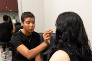 A woman with short hair is applying makeup to another woman with long hair in a salon. The makeup artist is holding a brush and seems focused on her work. There is a mirror and a cabinet in the background.