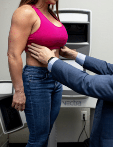 A woman in a pink sports bra and blue jeans is standing while a person in a blue suit examines her abdomen. The image appears to be taken in a medical or clinical setting with equipment labeled "VECTRA" in the background.