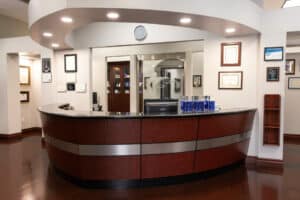 A modern office reception area with a curved, polished wooden desk and a computer monitor on top. Behind the desk is a glass wall with a suite number and a logo. The floor is shiny, dark wood, and the room is well-lit with ceiling lights. There are framed certificates and awards hanging on the walls. To the right, there is a wooden brochure holder mounted on the wall, and on the desk, there are multiple stacks of brochures.