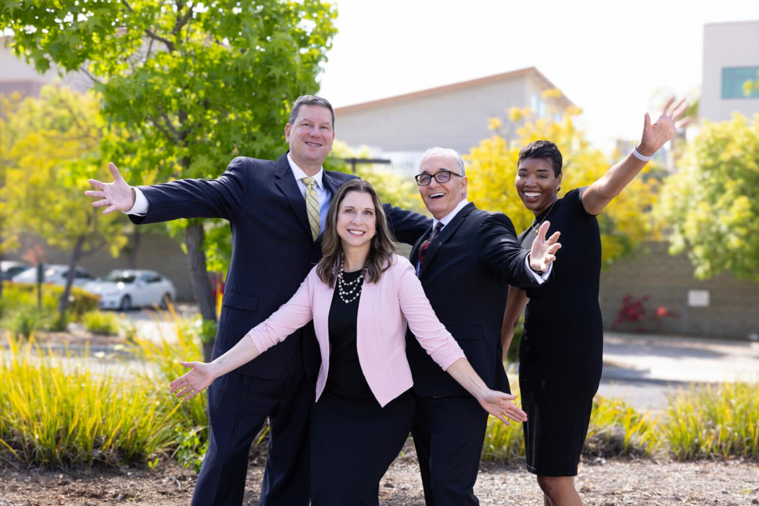 A group of four people dressed in business attire stand outdoors, smiling and posing with their arms outstretched. They appear cheerful and confident. The background shows trees, plants, and a building.