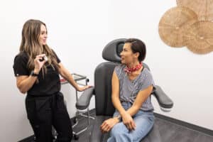 Two women are in a medical consultation room. One woman, dressed in black medical scrubs, is standing and speaking, while the other woman, dressed casually in a striped shirt and jeans, is sitting in a large chair, attentively listening. The room has a modern and clean aesthetic with neutral-colored walls and woven decorations on the wall.