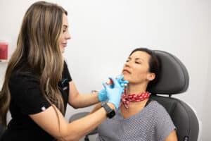A woman with long brown hair is administering an injection to another woman seated in a chair. The seated woman is wearing a striped shirt and a red scarf around her neck. The healthcare professional is wearing blue gloves and a black uniform. The setting appears to be a medical or clinic room.