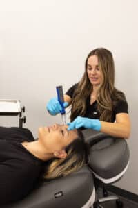 A woman wearing gloves is using a medical device on the forehead of another woman who is lying down on a padded chair in a clinical setting. The atmosphere appears professional and clean, with white walls and minimalistic decor.