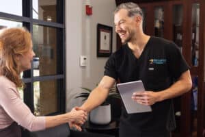 A man in black scrubs and holding a tablet is shaking hands with a woman inside an office. They are smiling at each other. The man has short gray hair and a beard, and the woman has long red hair. The office setting includes a framed certificate on the wall and a large window.