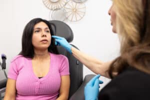 A woman with long dark hair, dressed in a pink ribbed shirt, is sitting on a medical examination chair. She appears to be in a consultation with a healthcare professional, who is wearing blue gloves. The healthcare professional is gently touching the woman's cheek, possibly discussing a medical or cosmetic procedure. The background includes some decorative items on the wall.
