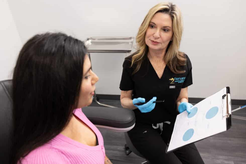 A woman in a black medical uniform and blue gloves is seated while holding a clipboard with charts. She is conversing with another woman who has long dark hair and is wearing a pink sweater. The setting appears to be a medical or consultation room.