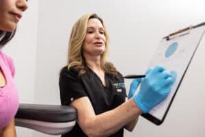 A healthcare professional wearing black scrubs and blue gloves is holding a clipboard and discussing it with a patient who is seated next to her. The patient has long dark hair and is wearing a pink shirt. They appear to be in a medical office setting.