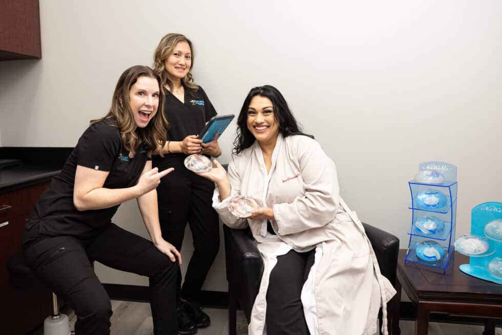 Three women in a medical office are smiling and engaging in a lighthearted manner. Two of them are wearing black medical uniforms, and one is holding a tablet while gesturing energetically. The third woman, seated and wearing a white medical coat, is holding a medical model. In the background, there are shelves displaying various medical models. The setting appears to be professional yet friendly.