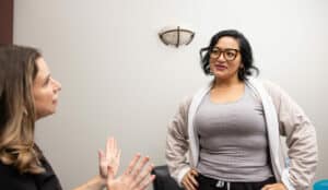 Two women are in conversation in an indoor setting. The woman on the left is engaged in the discussion, gesturing with her hands. She has long, wavy hair and is wearing a light-colored top. The woman on the right stands with her hands on her hips, wearing glasses, a gray tank top, and a light jacket. She seems to be listening attentively. The background has a plain wall with a light fixture.