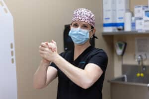 A healthcare worker wearing a surgical mask, black scrubs, and a pink leopard-print surgical cap is standing in a medical setting. They are rubbing their hands together, possibly as a hygiene or disinfecting procedure. The background includes medical supplies and equipment on shelves, and a sink with a faucet.