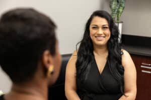 Two women are having a conversation in an indoor setting. The woman in the foreground, facing away from the camera, has short hair and is wearing earrings. The woman in the background has long, dark hair, a black sleeveless top, and is smiling warmly. There is a plant and office furniture visible in the background.