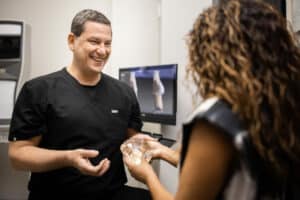 A healthcare professional in a black uniform is smiling and talking with a patient who has long, curly hair. The healthcare professional is holding clear anatomical models for demonstration. They appear to be in a medical office with medical equipment and a computer screen in the background.