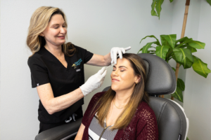 A medical professional wearing black scrubs and white gloves administers an injection to a woman's forehead. The woman receiving the injection is seated in a chair, eyes closed, and appears relaxed. There is a tall green plant in the background.