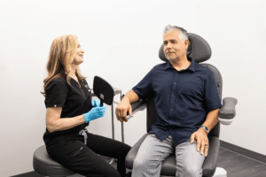 A healthcare professional wearing a black uniform and blue gloves is interacting with a seated patient in a clinical setting. The patient, who has gray hair and is wearing a navy blue polo shirt and gray pants, appears attentive and is seated in a chair designed for medical examinations. The room has white walls and minimal medical equipment visible.