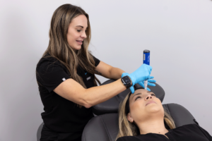 A woman wearing a black uniform and blue gloves is performing a cosmetic procedure on another woman who is lying down on a chair. The professional is holding a device to the forehead of the patient, who appears relaxed. The environment is clinical and clean, suggesting a medical or aesthetic treatment setting.