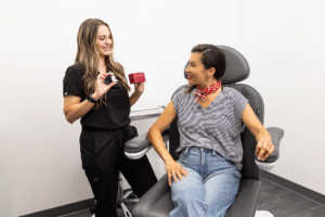 A woman sitting in a medical chair talking to a healthcare professional. The healthcare professional is holding a red box and smiling at the seated woman. The seated woman is wearing a striped shirt, jeans, and a red scarf around her neck. The healthcare professional is dressed in black medical scrubs. The setting appears to be a medical or consultation room with white walls.