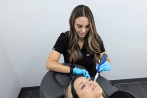 A woman in a black uniform and blue gloves performs a microneedling procedure on another woman lying back in a chair. The woman on the chair has her eyes closed and looks relaxed. The setting appears to be a clinical or spa environment, with a clean, professional ambiance.