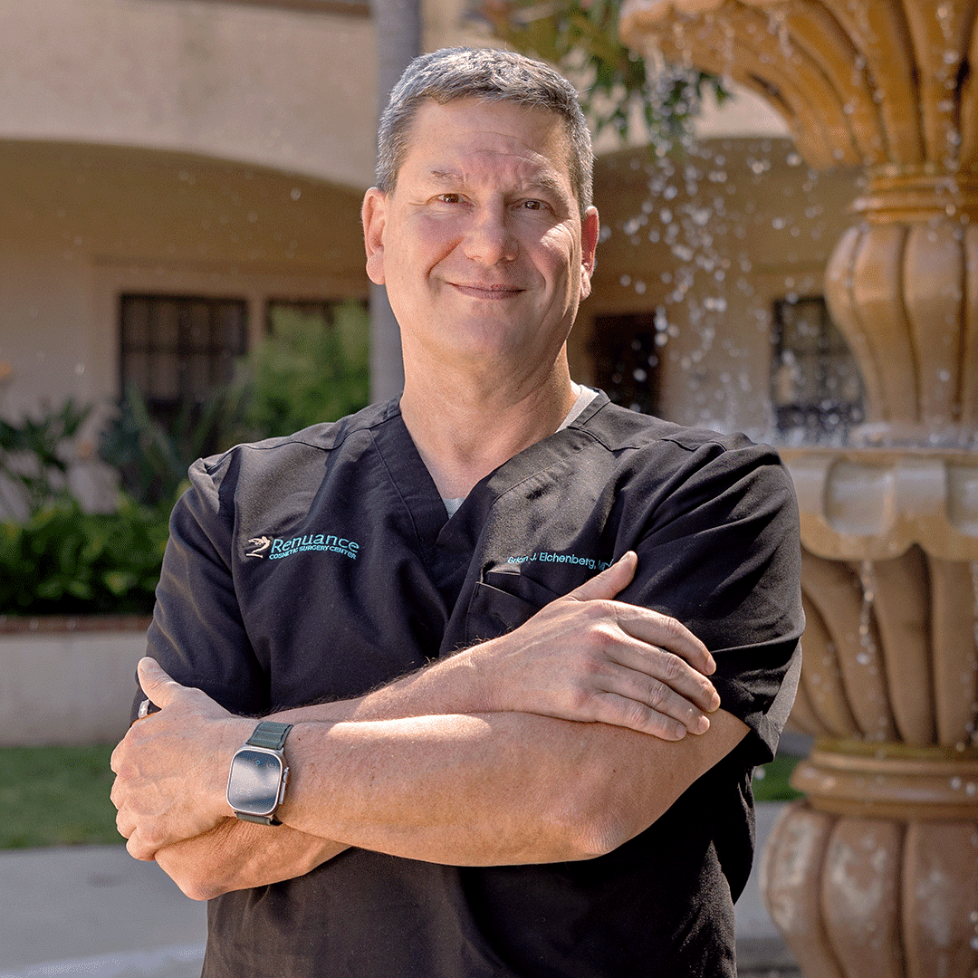 A smiling man stands outdoors in front of a fountain, with his arms crossed. He is wearing black scrubs with a logo and name embroidered on the chest, as well as an Apple Watch on his left wrist. The background features a building with windows and lush green plants.