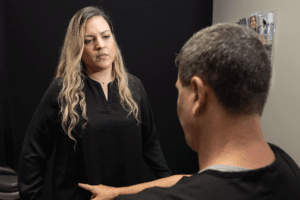 Two people are engaging in a conversation in an indoor setting. The person on the left has long, wavy blond hair and is wearing a black top. The person on the right, whose back is to the camera, has short gray hair and is wearing a black shirt. The background appears to be a dark, nondescript interior. The interaction seems serious or focused.