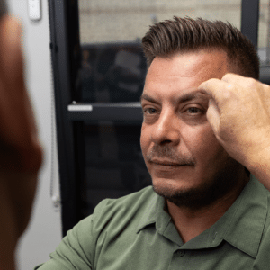 A person in a green shirt is sitting while another person in the foreground, blurred, is using their hand to touch the first person's right eyebrow. They are indoors, and there is a window in the background.