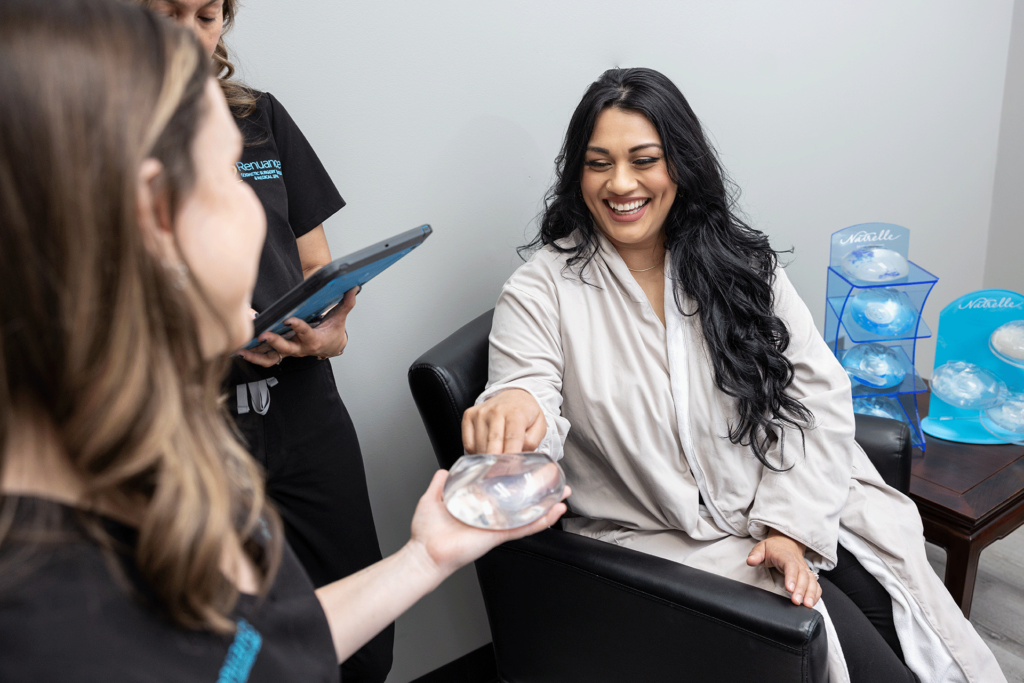 A woman with long dark hair is sitting in a black chair and smiling while holding a breast implant sample handed to her by a healthcare provider. She is wearing a light-colored robe. Another healthcare provider is standing in the background, holding a tablet and observing. The setting appears to be a medical consultation room, with additional breast implant samples displayed on a table to the side.