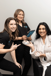 Three women are pictured in a clinical setting. The woman seated on the right is dressed in a white robe and is smiling while holding translucent objects that appear to be breast implants. Two other women on the left, both dressed in black scrubs, are standing and smiling. One is pointing towards the objects while the other is holding a tablet. The background is a plain, light-colored wall.