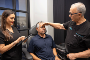An older male doctor is examining the forehead of a seated male patient in a medical office. A female medical assistant stands to the side, holding a tablet and taking notes. The medical team is dressed in black uniforms. The patient wears a dark blue shirt and appears attentive. The room has a modern design with a window and a chair for the patient.