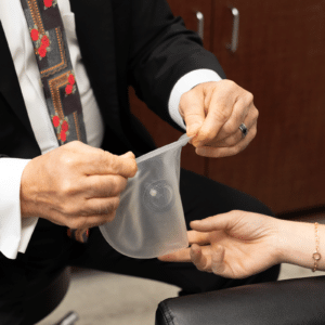 A person in a suit jacket and decorative tie is demonstrating how to use a female condom by stretching it open. Another person's hand is visible in the foreground, reaching out towards the condom. The setting appears to be an office or a professional environment.