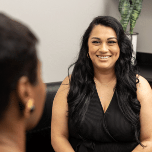 A woman with long black hair smiles while sitting and facing another person with short hair in a professional setting. She is wearing a black sleeveless top and a necklace, and there is a green leafy plant in the background.