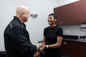 A man and a woman shaking hands in an office setting. The woman, dressed in a black top, is smiling warmly, while the man, wearing a dark jacket, also appears to be smiling. The office has minimalist decor, with a few cabinets, a small sink, and a wall-mounted light.