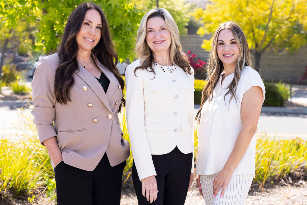 Three women standing outdoors, smiling at the camera. The woman on the left is wearing a beige blazer and black pants, the woman in the middle is wearing a white blazer and black pants, and the woman on the right is wearing a white blouse and light-colored pants. They are standing in front of a green, leafy background with trees and bushes, and the sun is shining brightly.
