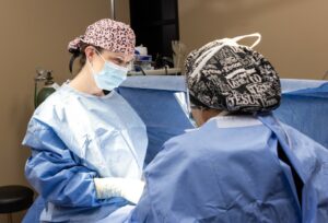 Two surgeons in an operating room performing a surgery. One surgeon is facing the camera, wearing a pink patterned surgical cap, a face mask, and blue surgical attire. The other surgeon has their back to the camera and is wearing a black and white patterned surgical cap with text, a face mask, and blue surgical attire. Medical equipment is visible in the background.